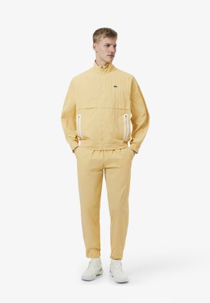 Homme debout portant un survêtement jaune avec des poches zippées et des baskets blanches, regardant sur le côté devant un fond uni.