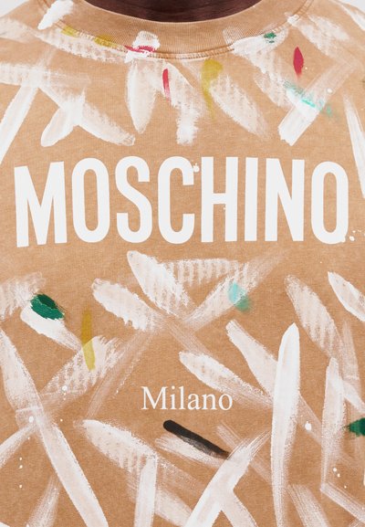 Μπεζ πουκάμισο με λευκά πινέλα και πολύχρωμες πιτσιλιές μπογιάς, με έντονα λευκά γράμματα "MOSCHINO" και μικρότερα "Milano".
