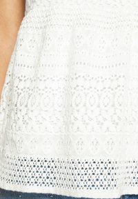 Haut blanc à motifs avec de la dentelle texturée, dotée de designs géométriques et de détails en tissage ouvert, terminé par un ourlet uni.