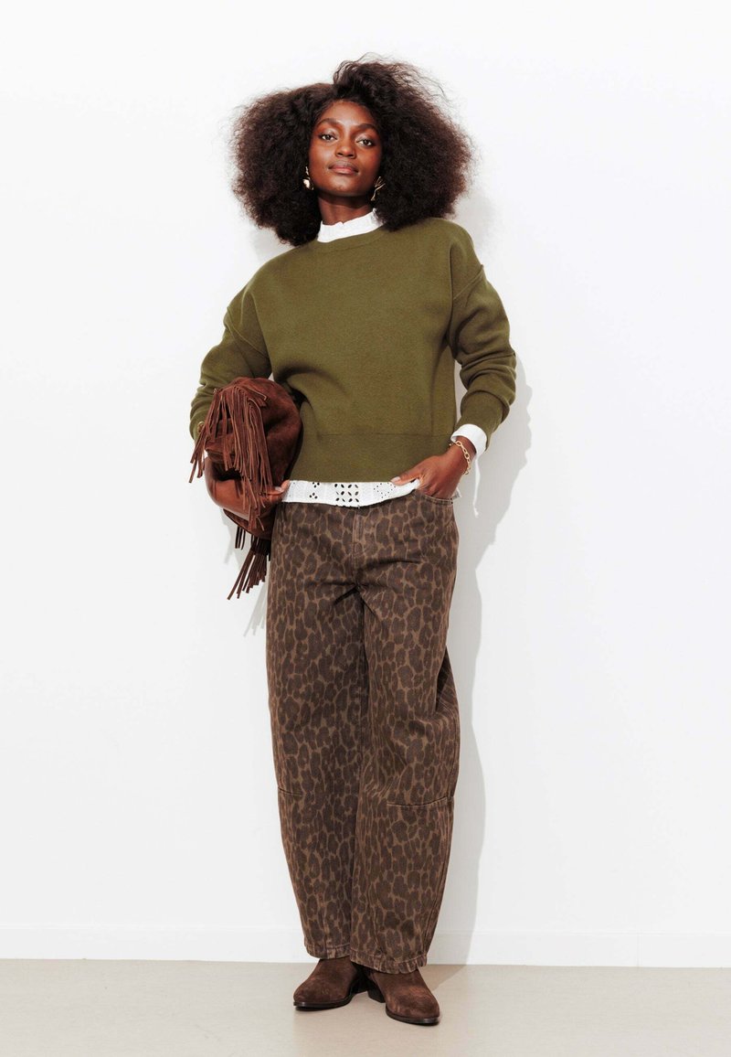 Femme aux cheveux bouclés portant un pull vert olive, un chemisier blanc, un pantalon à imprimé léopard, tenant un sac à franges marron, debout contre un mur blanc.