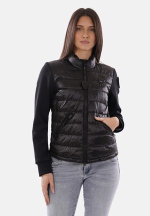 Donna che indossa una giacca nera trapuntata con cerniera e tasche, in piedi con una mano nella tasca e jeans azzurri chiari su uno sfondo semplice.