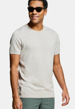 Man met zwarte zonnebril, lichtbeige T-shirt met korte mouwen en groene broek, glimlachend voor een effen witte achtergrond.
