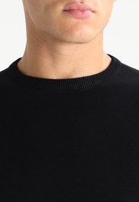 Schwarzer Strickpullover mit Rundhalsausschnitt, glatter Textur und Rippdetails am Kragen. Nahaufnahme des Halsbereichs.