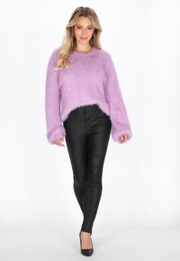 Donna che indossa un maglione fuzzy lavanda con dettagli in perle, pantaloni neri aderenti e tacchi neri, in piedi contro uno sfondo bianco.