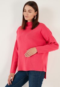 Pull long à manches longues rose avec un col haut ; texture côtelée, coupe ample et fentes latérales, associé à un jean en denim foncé.