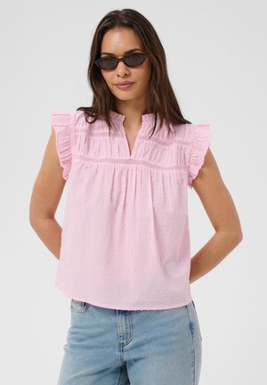 Frau mit schwarzen ovalen Sonnenbrillen und einer ärmellosen pinken Bluse mit Rüschen sowie hellblauen Jeans, vor neutralem Hintergrund stehend.