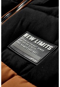 Czarna i brązowa kurtka puchowa z teksturowaną czarną gumową naszywką z wytłoczonym białym tekstem i akcentami w postaci pasków.