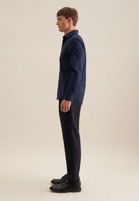 Mann im Profil stehend, trägt ein dunkelblaues Langarmhemd, schwarze Hose und schwarze Anzugschuhe vor einem schlichten hellen Hintergrund.