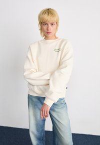 Jong persoon met kort blond haar draagt een oversized crèmekleurig sweatshirt en vervagde blauwe jeans, staande tegen een effen witte achtergrond.