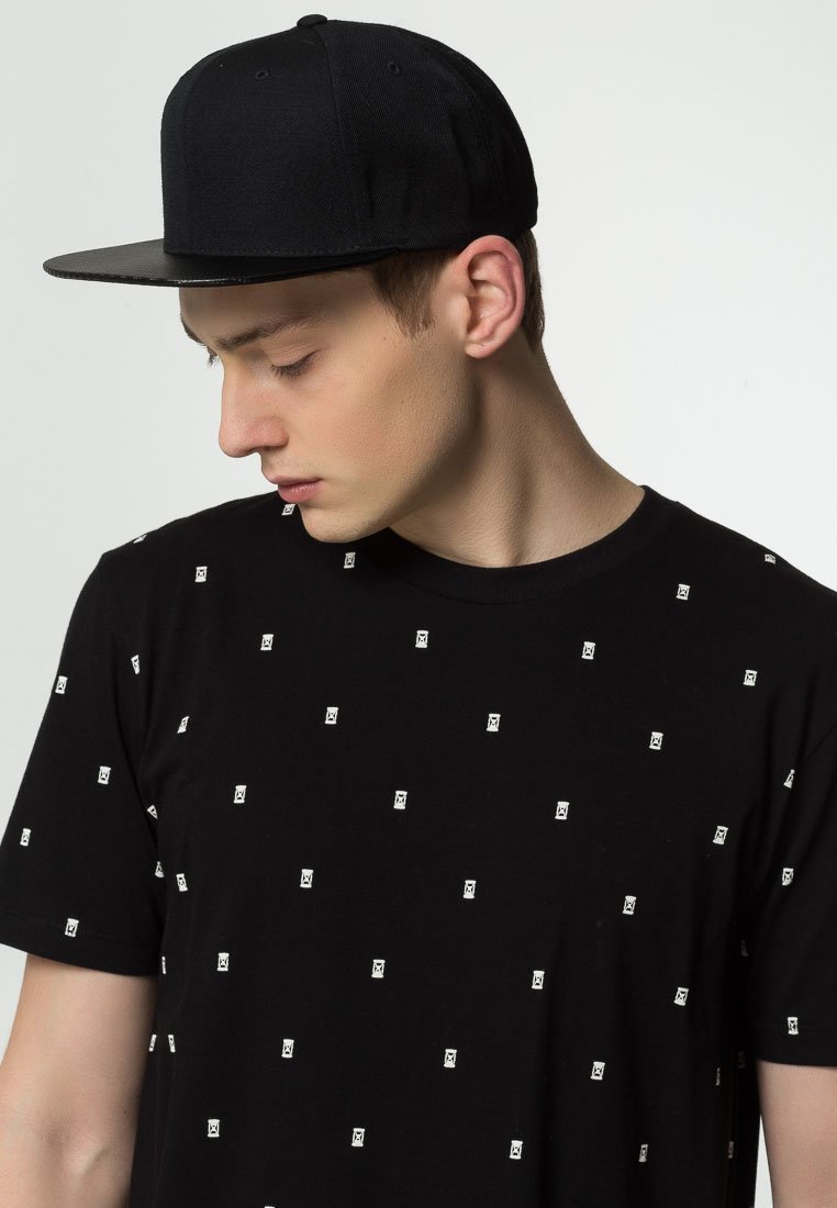 Căciulă neagră cu o bordură plată, purtată cu un tricou negru ce prezintă un model mic de pătrate albe. Materialul pare neted, cu mâneci scurte.
