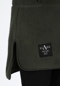 Olijfgroene fleece kleding met zijslitsen, contrasterend zwart label, zachte textuur en afgeronde randen. Ontworpen voor comfort en warmte.
