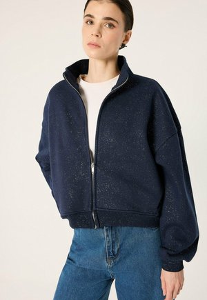 Jeune femme portant une veste zippée bleu marine à pois par-dessus une chemise blanche et un jean bleu, debout devant un fond clair uni.