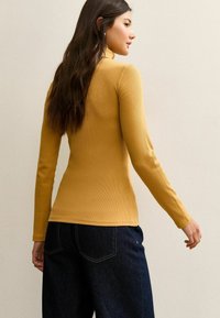 Maglia a maniche lunghe color senape a coste con design aderente. La parte posteriore presenta un collo a girocollo e una texture liscia, abbinata a jeans scuri in denim.