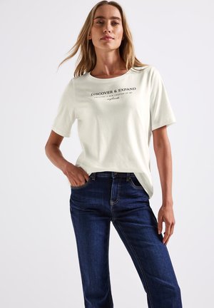 T-shirt en coton de couleur claire à manches courtes, avec un texte noir sur le devant. Associe à un jean en denim bleu foncé.