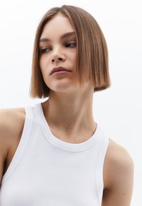 Vit ribbad linne med breda axelremmar och rund halsringning, placerad mot en enfärgad bakgrund.