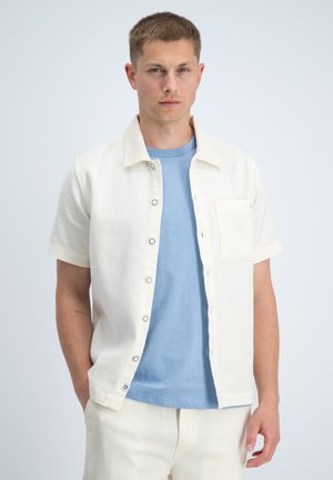 Hombre que lleva una camisa blanca de manga corta abotonada sobre una camiseta azul claro, con pantalones blancos, de pie contra un fondo liso y claro.