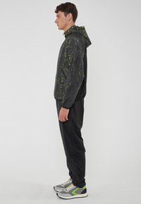 Giacca a vento da uomo nera con motivi astratti verdi, abbinata a pantaloni neri e sneakers grigie con accenti verdi. Tessuto liscio.