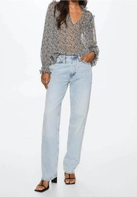 Femme portant un chemisier transparent à motifs noir et blanc avec des poignets smockés, un jean droit taille haute bleu clair et des sandales à talons noires.