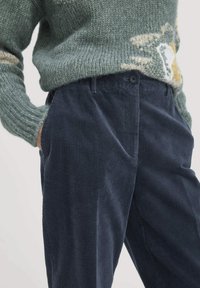 Marineblauwe corduroy broek met verticale ribbels, voorzien van voorzakken en een knoopsluiting, gecombineerd met een gestructureerde groene gebreide trui.