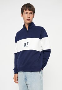 Námornícky modrý a biely fleecový pulover s kvartovým zipsom, ktorý v strede zdobí logo "GAP". Mäkká textúra a voľný strih.
