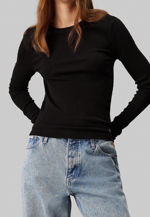 Kobieta ubrana w dopasowaną czarną bluzkę z długim rękawem, wpuszczoną w wysokouwęższe jasnoniebieskie dżinsy, stojąca z rękami na biodrach.