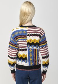Pull en tricot multicolore présentant des motifs géométriques audacieux, avec des rayures bleu, jaune et bordeaux ; ourlet et poignets à côtes noirs.