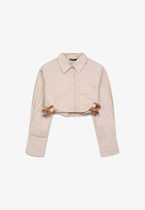 Chemise beige raccourcie à manches longues, col à boutonnière, poche avant, et sangles en cuir à l'ourlet pour un ajustement réglable. Texture de tissu lisse.