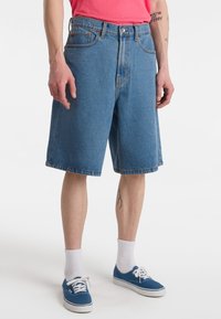 Szorty dżinsowe w średnim odcieniu niebieskiego, do kolan, z postrzępionym brzegiem, o kroju z pięcioma kieszeniami, zapinane na metalowy guzik. Białe skarpetki i niebieskie trampki.