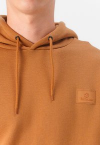 Sweatshirt à capuche marron en tissu doux, avec un patch logo texturé sur la poitrine gauche et des cordons de serrage réglables à la capuche.