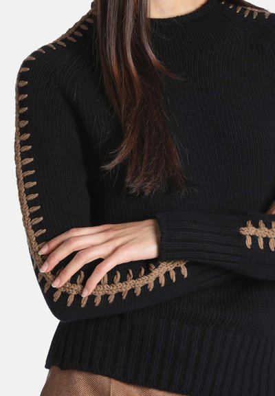 Pull noir en maille avec des détails marron cousus le long des manches et des épaules, porté par une personne aux bras croisés.
