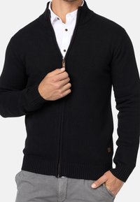 Maglione nero lavorato a maglia con colletto e polsini a coste, chiusura con zip e piccolo etichetta del marchio vicino all'orlo. Indossato sopra una camicia bianca con colletto.