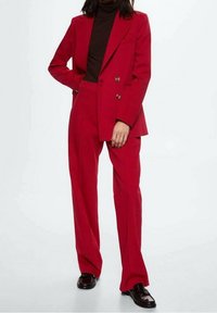 Femme portant un costume-pantalon rouge vif avec une veste croisée et un pantalon à jambes larges, associé à un col roulé noir et des mocassins foncés.