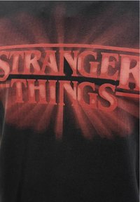 Must t-särk, millel on julgelt punased tähed "STRANGER THINGS" gradiendi efektiga, tumeda, hääbuv taust.