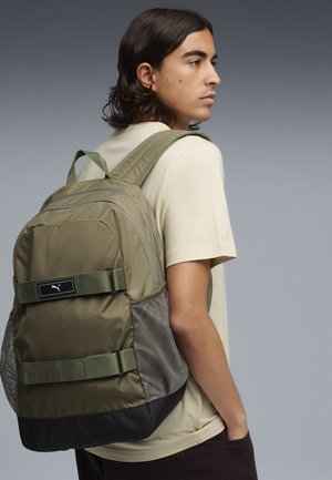 Joven con cabello de longitud media, vistiendo una camisa beige y una mochila Puma verde oliva, de pie de lado contra un fondo gris liso.