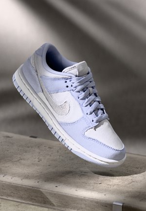 Niskie sneakersy w białym i jasnoniebieskim kolorze z teksturowanym materiałem i sznurowadłami, pokazane unoszące się nad betonową powierzchnią.