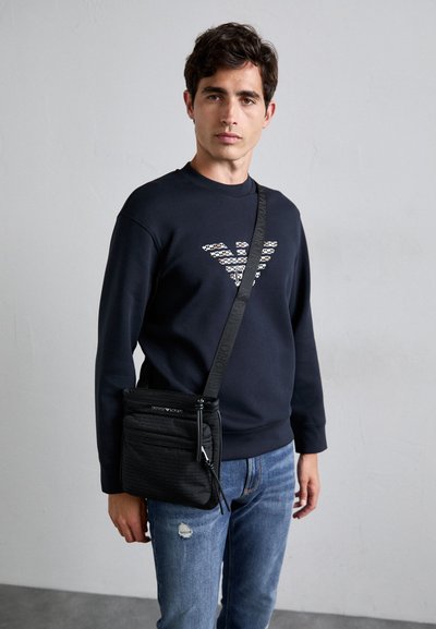 Bolso bandolera negro con tejido texturizado, bolsillo delantero con cremallera y correa con logo. Combinado con un sudadera azul marina y jeans desgastados.