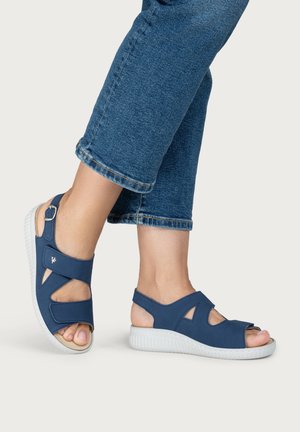 Person trägt blaue offene Sandalen mit verstellbaren Riemen und weißen Sohlen, kombiniert mit gekürzten Jeans.