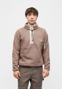 Νεαρός άνδρας που φοράει καφέ φλις πουλόβερ με ψηλό γιακά και τσέπη στο στήθος με φερμουάρ, στέκεται μπροστά σε απλό λευκό φόντο.