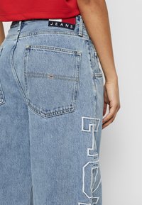 Světle modré džíny mají vysoký pas, zadní kapsy a velká bílá vyšitá písmena na boku. Štítek zní "JEANS."