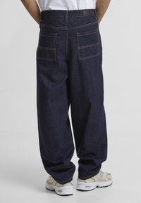 Pantalones de mezclilla oscura con un ajuste holgado, que cuentan con dos bolsillos traseros y costuras amarillas en contraste. Se llevan con zapatillas de color claro.