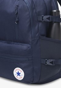 Marinblå ryggsäck i hållbart tyg, med en dragkedjeficka, nätficka på sidan för en vattenflaska och en rund Converse All Star-logga.