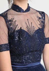 Abito blu navy con un corpetto trasparente ricamato che presenta paillettes e motivi floreali. La cintura in raso e le maniche aderenti migliorano il design.