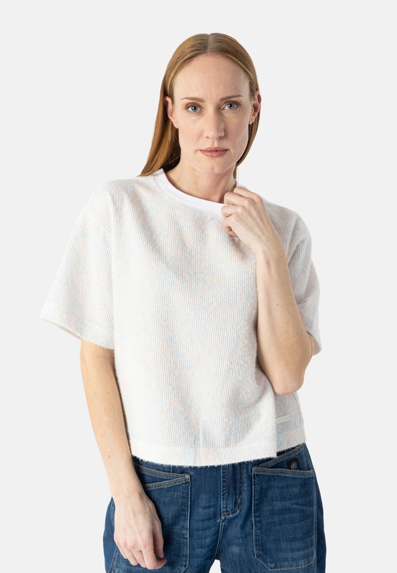 Femme aux cheveux blonds raides portant un pull blanc côtelé à manches courtes et un jean bleu, regardant devant elle avec une expression neutre.