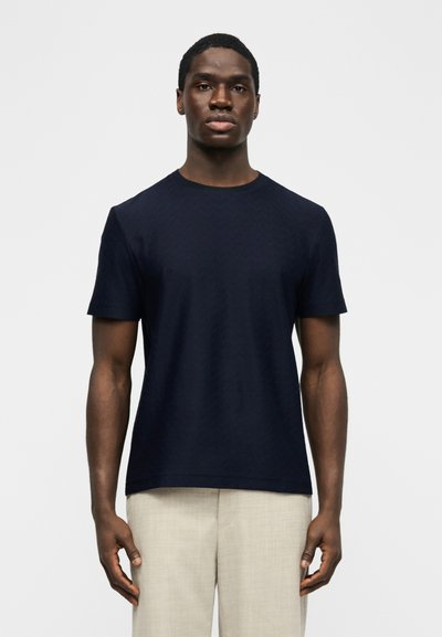 Άνδρας που στέκεται μπροστά σε απλό φόντο, φορώντας μπλε σκούρο κοντομάνικο μπλουζάκι και ανοιχτό μπεζ παντελόνι.