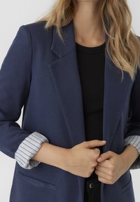 Mörkblå kavaj med en slagkrage, försedd med randiga detaljer vid ärmsluten och en slät textur. Matchad med en svart ribbad topp.