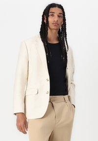 Beige blazer met twee gouden knopen, gestructureerde stof, gekleed met notched lapels en twee zijzakken, gedragen over een zwart shirt en beige broeken.