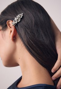 Silberner Haarclip mit einem Blattdesign und markisenförmigen Steinen. Trägt sich auf dunklem Haar und hebt die filigranen Details und den Glanz hervor.