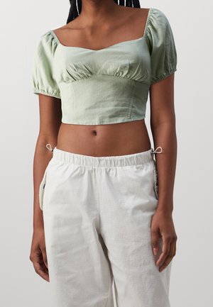 Vrouw draagt een lichtgroene cropped top met pofmouwen en witte, hooggetailleerde broek met koordsluitingen aan de zijkanten.