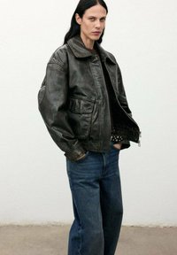 Chaqueta de cuero marrón oscuro con un cuello ancho y dos bolsillos frontales, combinada con unos vaqueros azules holgados, sobre un fondo blanco simple.