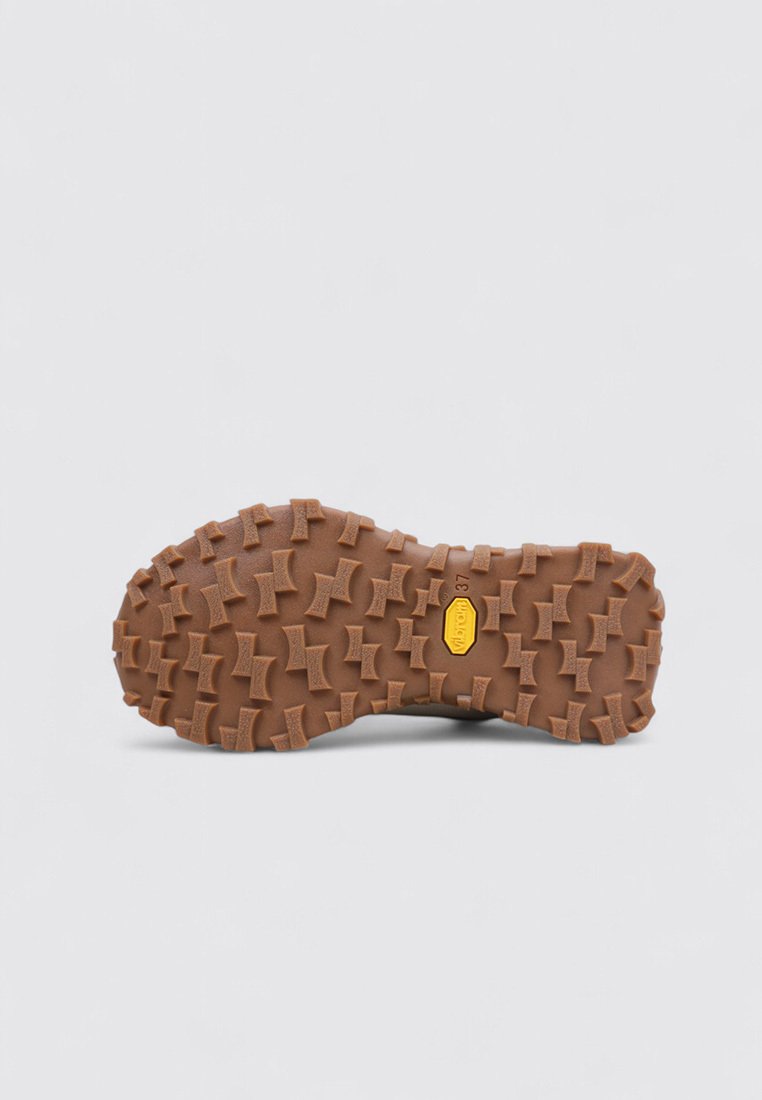 Rubberen zool met diepe, robuuste loopprofiel in bruin, voorzien van een levendig geel logo en een gestructureerd oppervlak voor verbeterde grip.